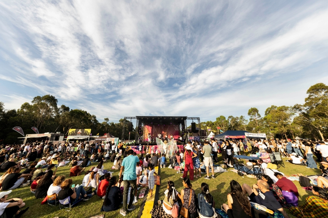 A large crowd gathers on a grassy field to enjoy an outdoor concert under a partly cloudy sky. The red stage is vibrant, and the scene is lively and cheerful.