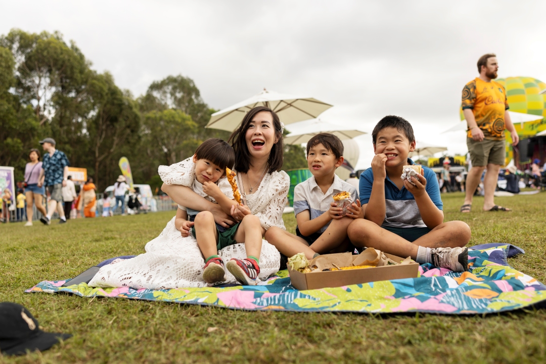 A joyful family picnic: a woman and three children sit on a colourful blanket in a park. The sky is cloudy, and other people are gathered in the background.