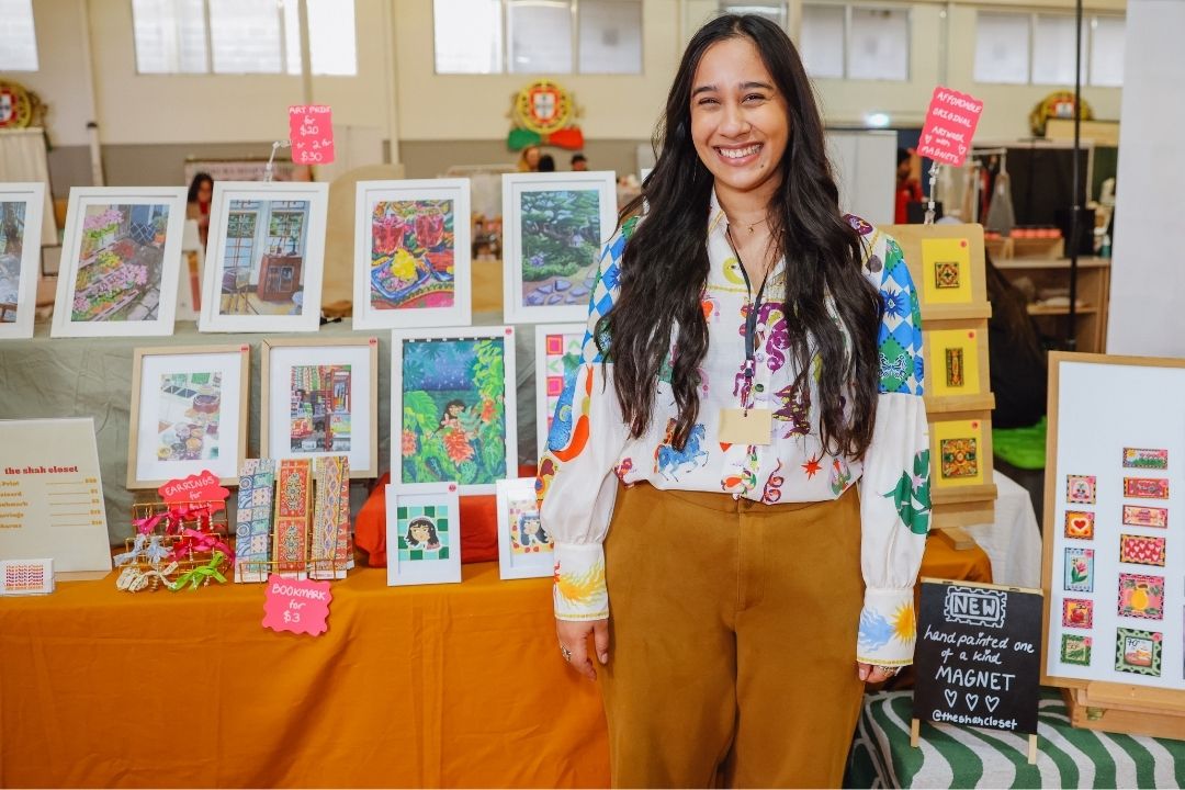 A woman smiles at a craft fair booth displaying colourful art prints and creative items. The setting is bright and lively, evoking a cheerful atmosphere.