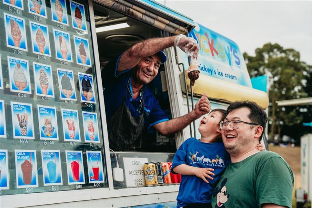 A man serves ice cream from a truck to two smiling customers—a man and child, capturing a joyful interaction on a sunny day. Ice cream menu visible.