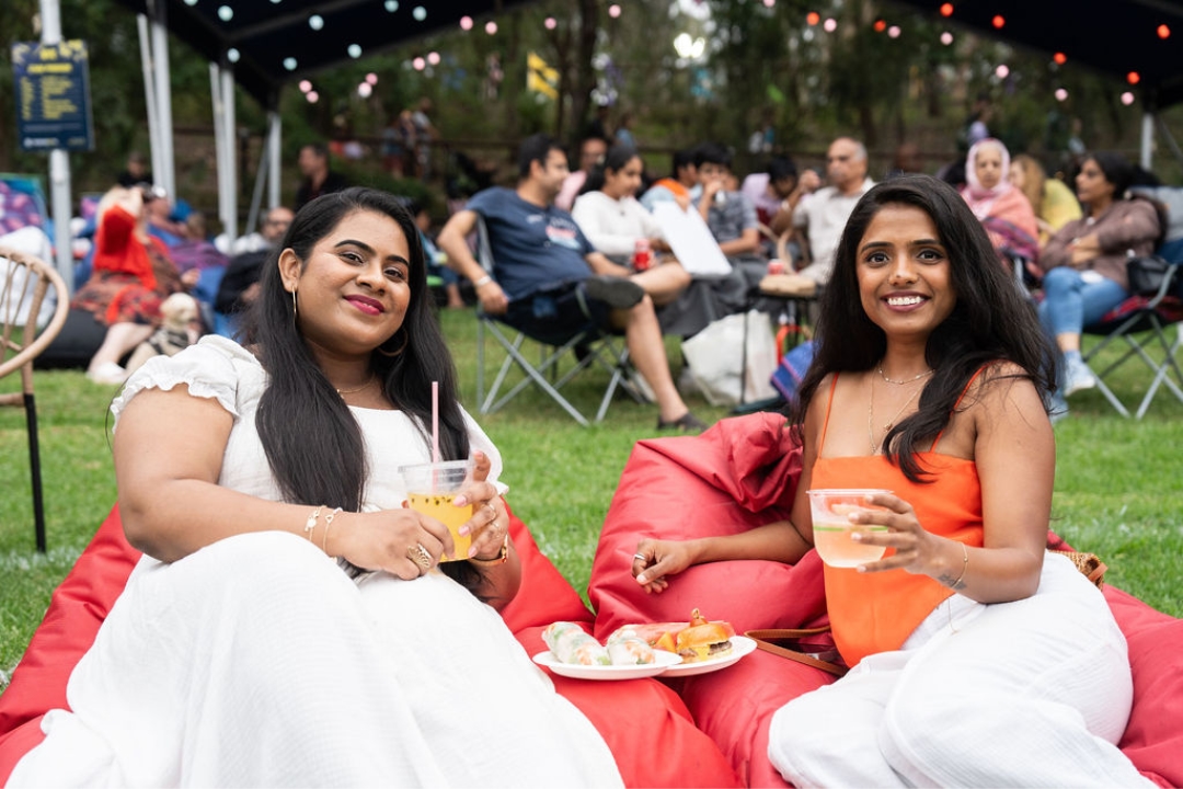 Two women smiling, seated on red bean bags at an outdoor event, holding drinks. A picnic scene with a relaxed crowd in the background under string lights.