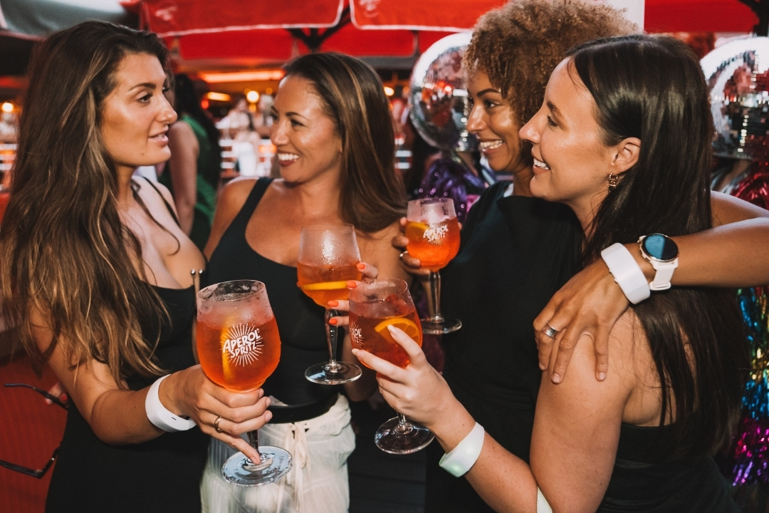 Four women in stylish attire enjoy a festive gathering, holding orange drinks and smiling warmly in a lively, colourful party atmosphere.