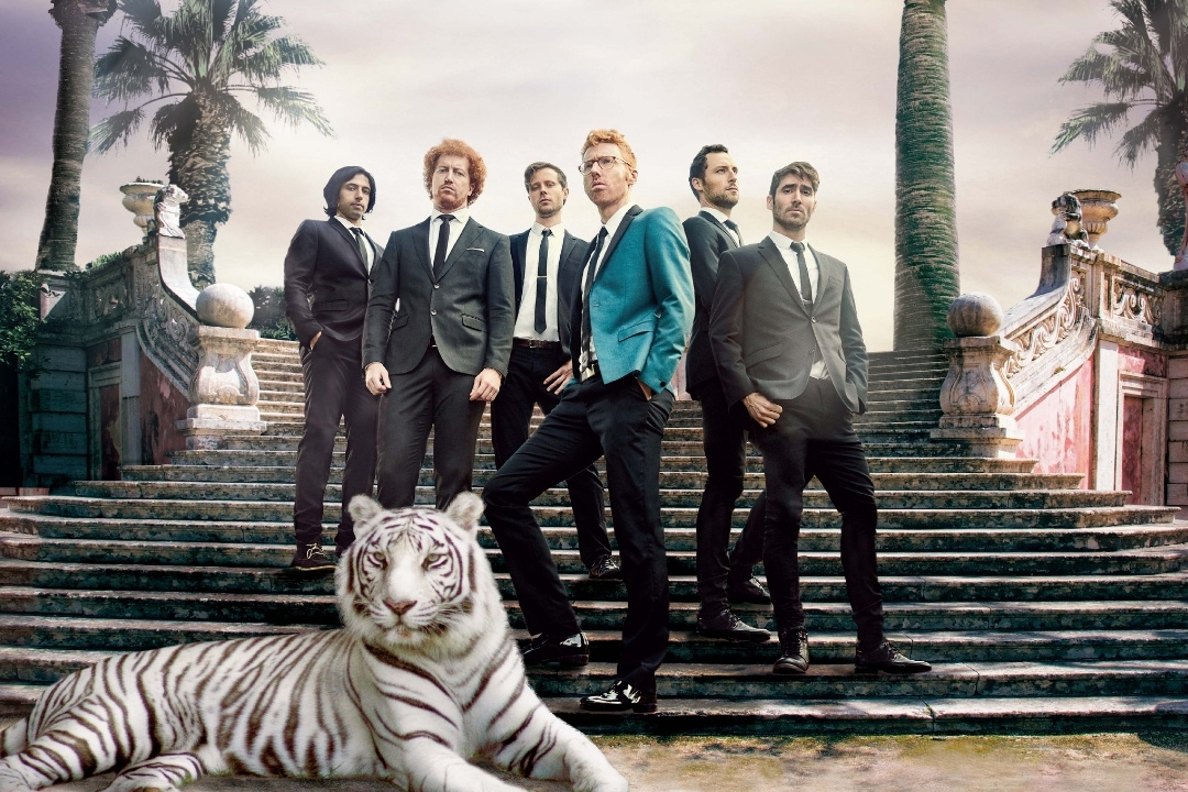 A group of five men in formal suits stands confidently on stairs flanked by palm trees. A white tiger reclines in the foreground, adding a surreal touch.