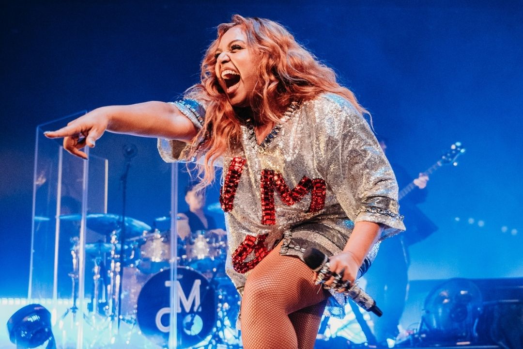 A vocalist energetically performs on stage, pointing forward with a microphone. Blue stage lights create an exciting and dynamic concert atmosphere.