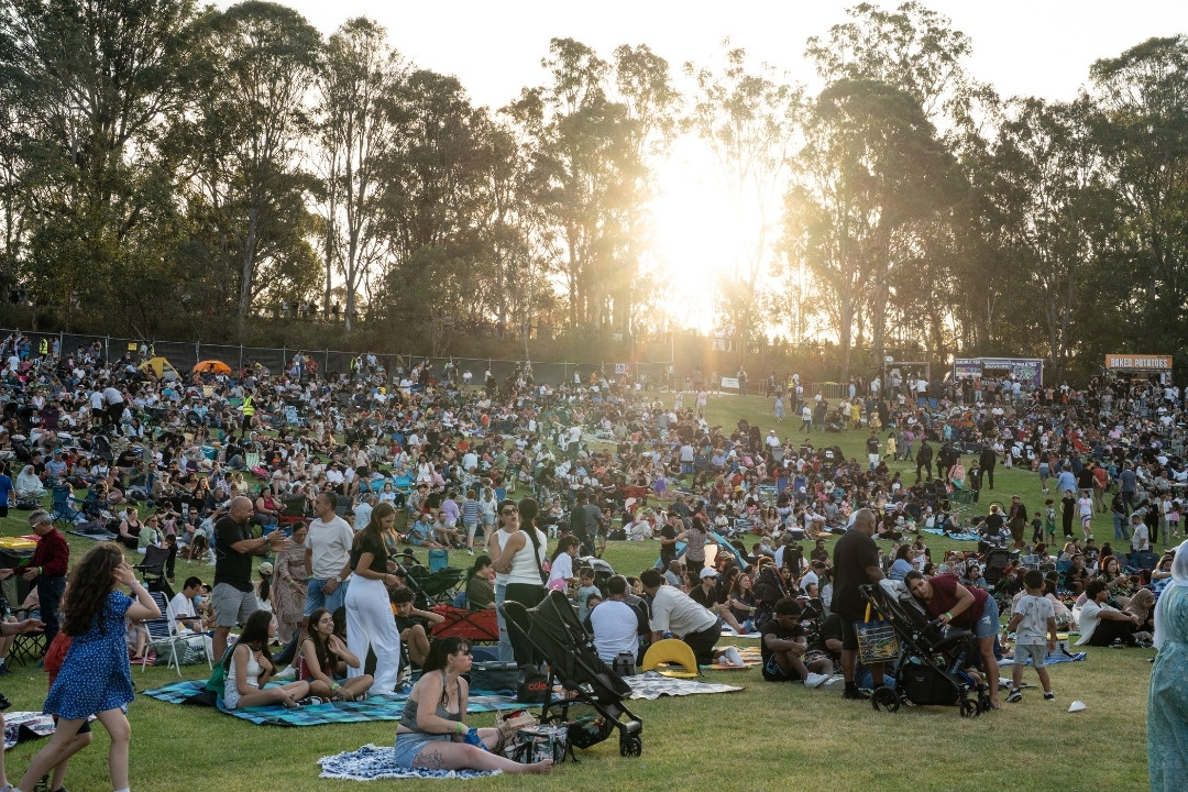 A large crowd sits on a grassy field at sunset, surrounded by tall trees, enjoying an outdoor event. The atmosphere is relaxed and social.