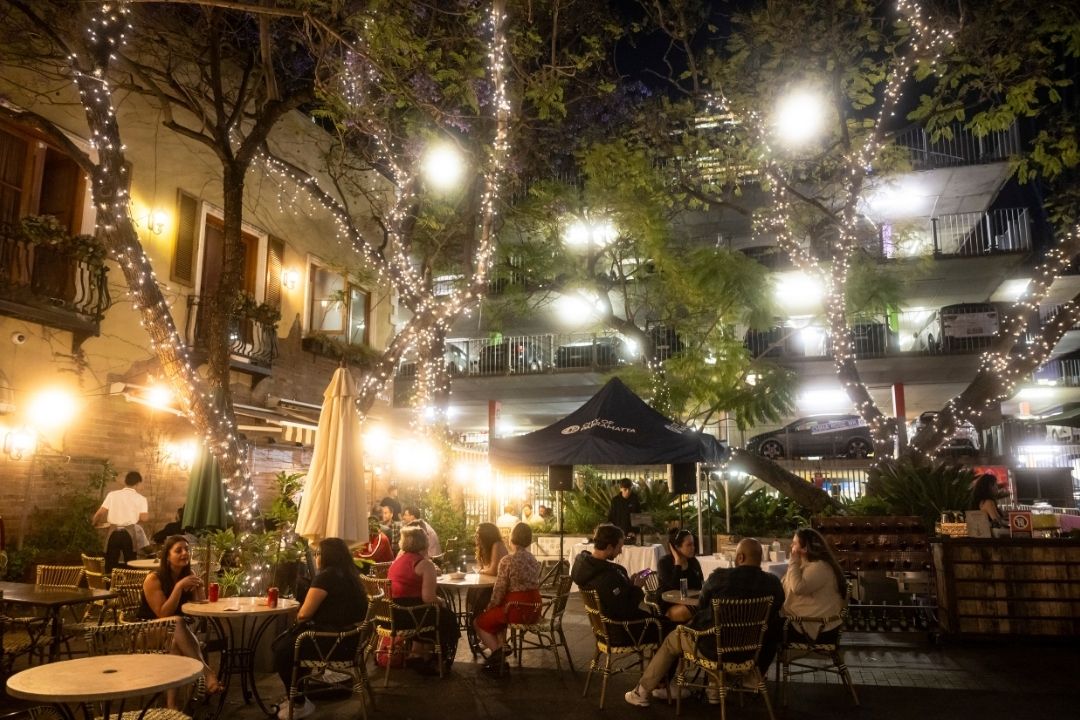 Cozy outdoor café at night with string lights illuminating tables and people dining under trees. The scene is warm and inviting with a lively atmosphere.