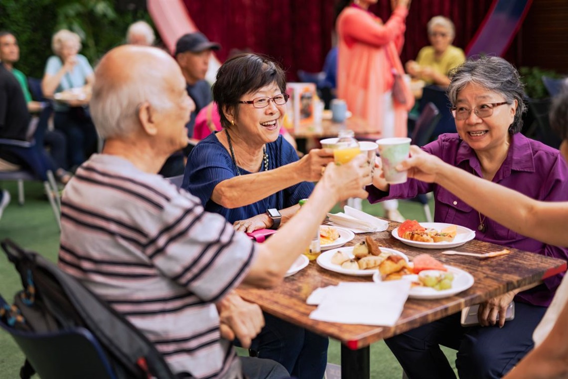 A group of older adults sitting at a table clinking glasses in celebration. They are smiling, enjoying food, and surrounded by a festive atmosphere.
