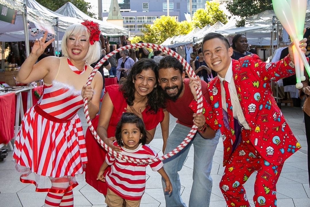 A joyful group posing together at a festive outdoor event. Adults wear red and white outfits, while a child in stripes grins, holding a hoop.