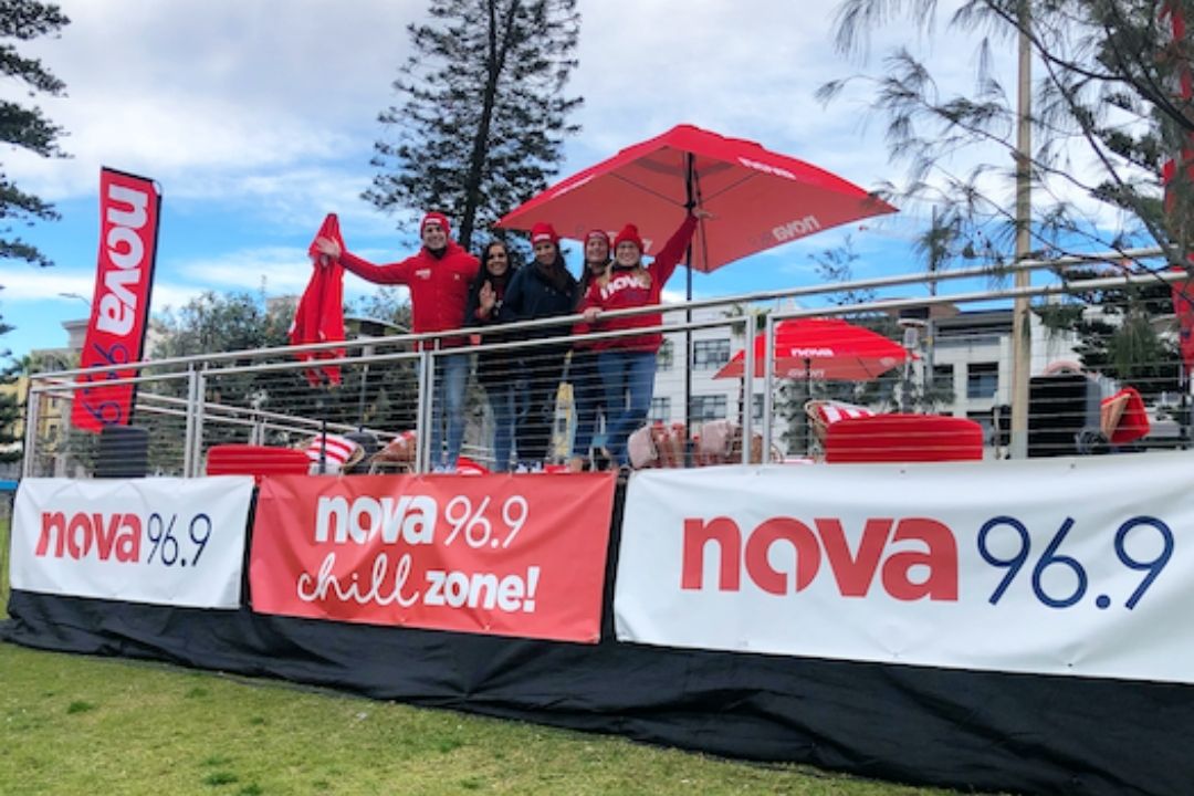 Outdoor event booth with "Nova 96.9" branding. Staff in red shirts engage with attendees. Bright banners and flags create a lively, festive atmosphere.