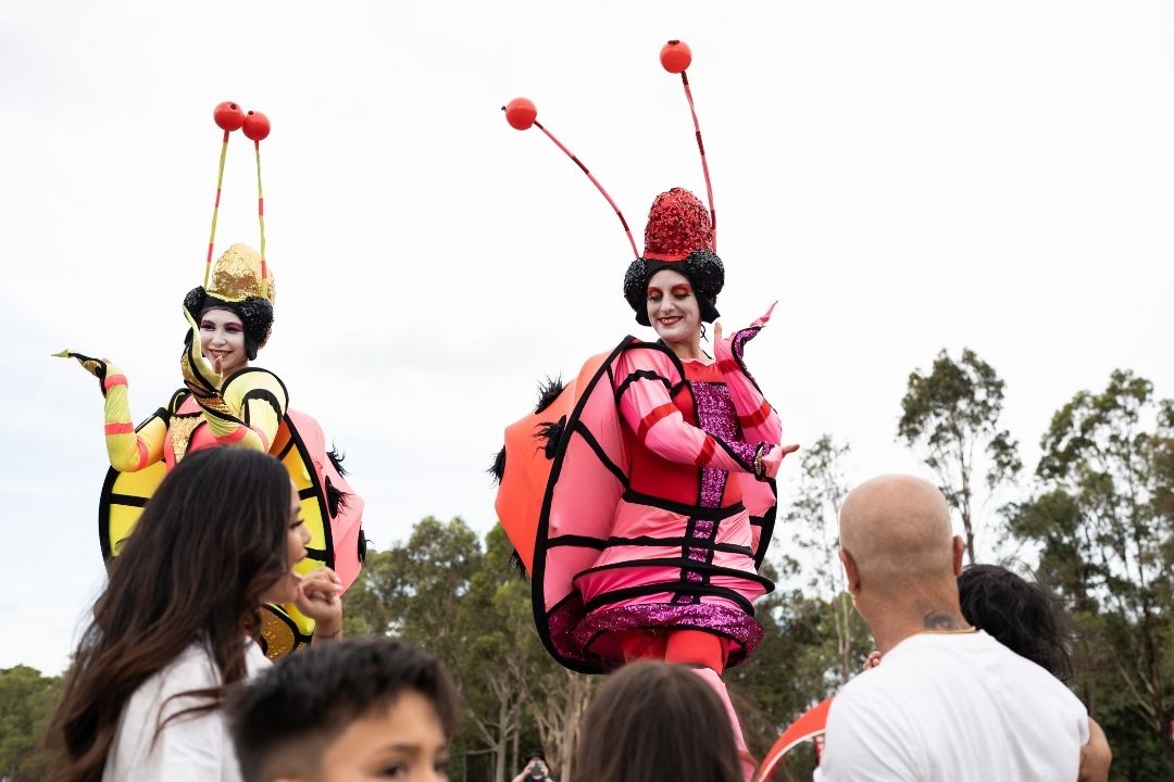 Two performers in colourful insect costumes, one yellow and one pink, entertain a crowd outdoors. They are smiling, creating a joyful and lively atmosphere.