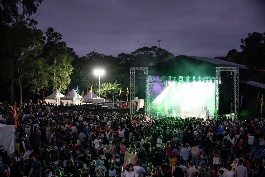 Outdoor concert at night with a large crowd gathered in front of a brightly lit stage. Trees border the venue, and the atmosphere is lively and energetic.