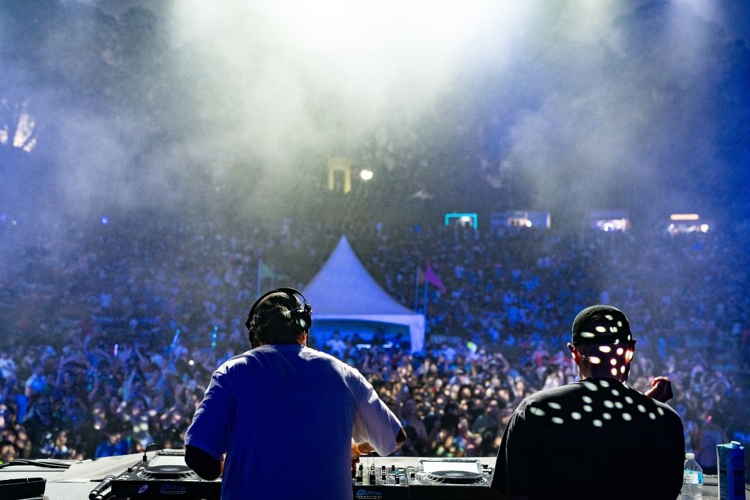 Two DJs perform on stage at a lively outdoor concert, with a large crowd and bright lights creating an energetic, electrifying atmosphere.