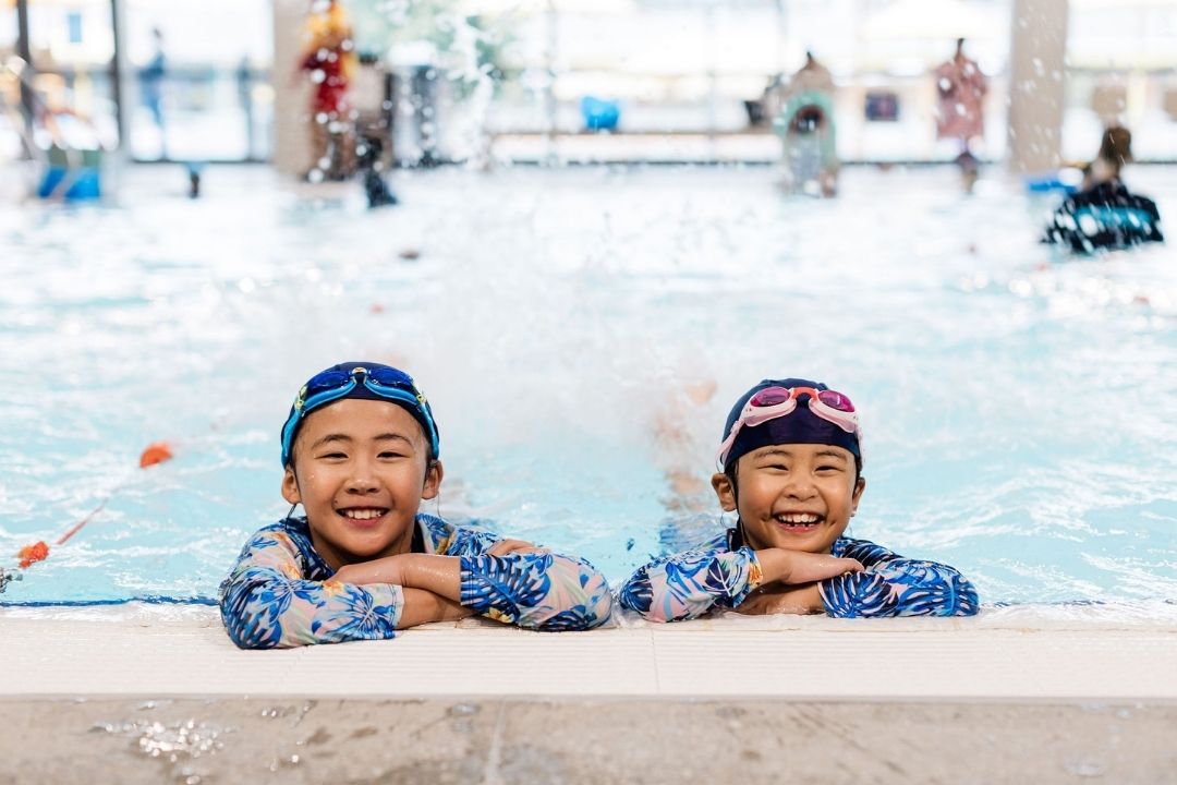 Two children in colourful swimwear and goggles joyfully lean on the edge of a pool. Splashes of water surround them, highlighting their fun swim session.