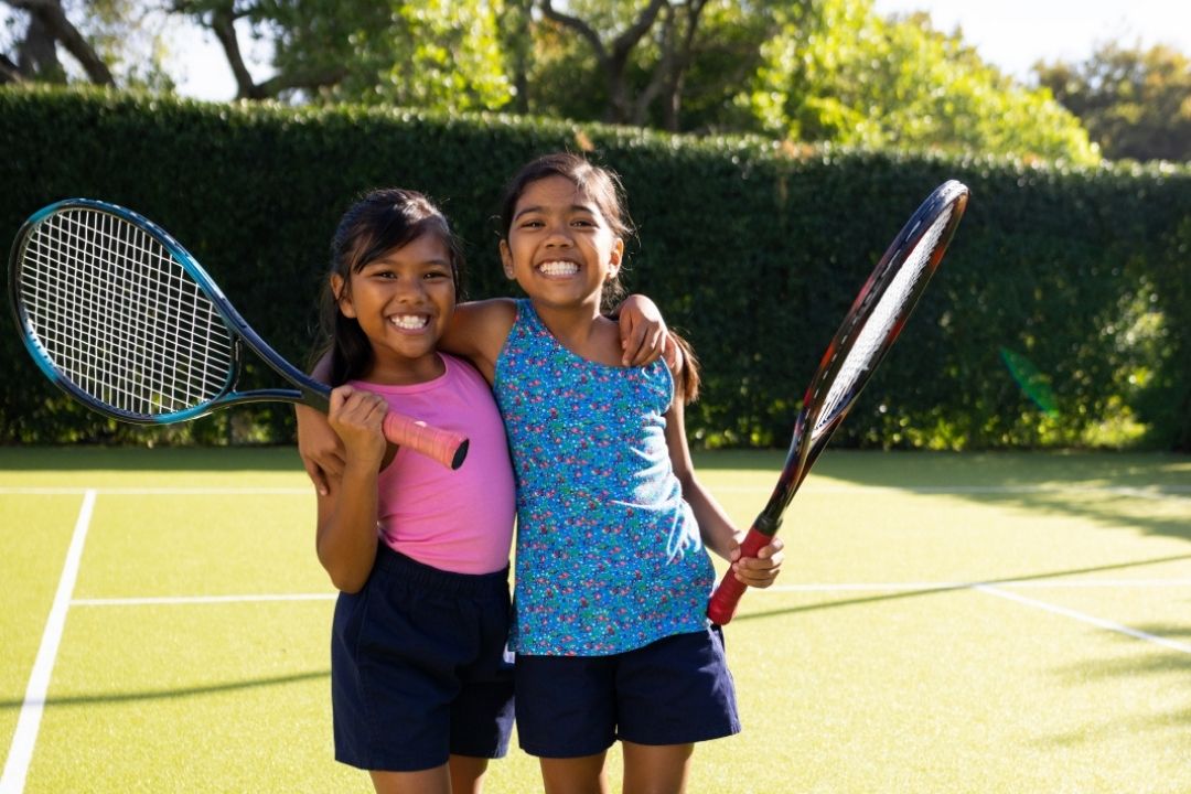 Two smiling girls stand on a sunny tennis court, each holding a racket. They're embracing, dressed in sporty outfits, with trees in the background.