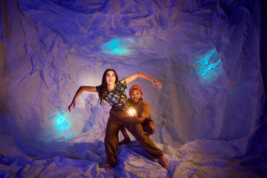 A woman and a creature with four arms and glowing blue orbs stand in a mystical cave with a purple hue. The scene conveys a sense of fantasy and adventure.