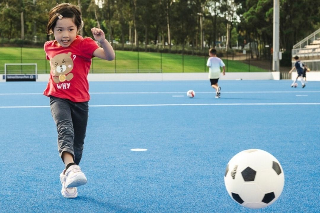A child in a red shirt joyfully runs on a blue sports field, chasing a soccer ball. Another child plays in the background; the scene is lively and playful.