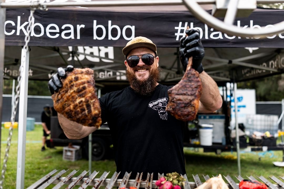 A bearded man wearing sunglasses and a cap stands smiling at an outdoor barbecue event, holding two large, grilled meats in front of a "Blackbear BBQ" tent.
