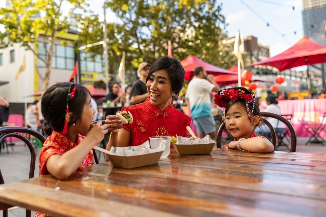 A woman and two children sit at an outdoor table, enjoying dessert waffles. They appear joyful and engaged, with bright umbrellas and greenery in the background.