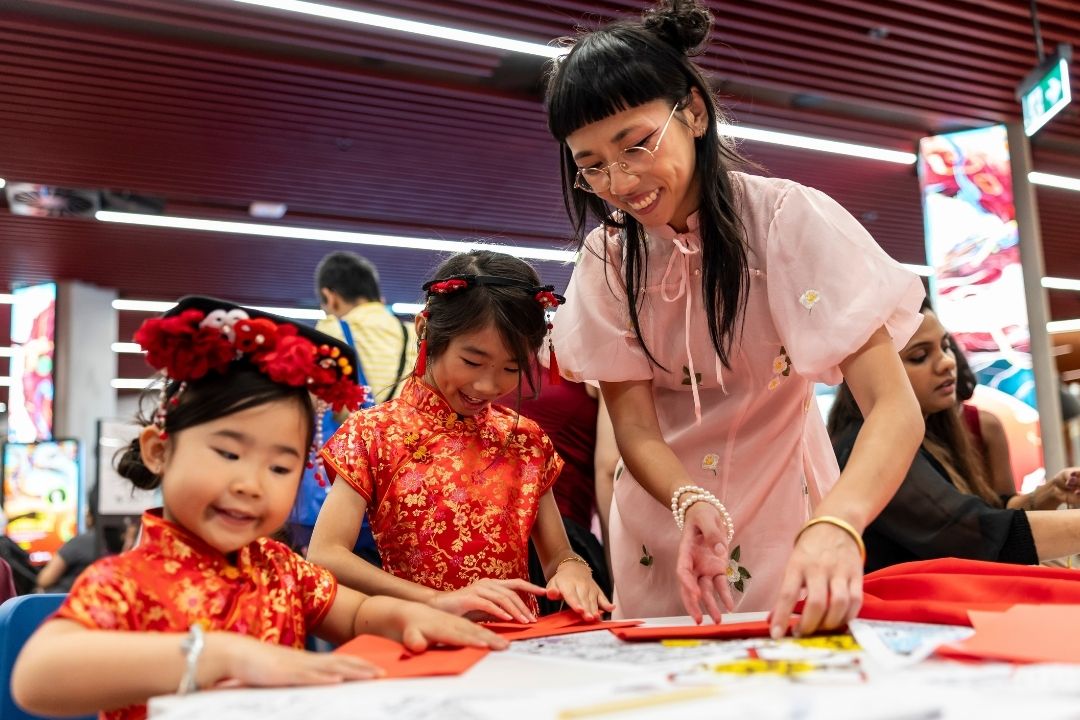A woman and two children, wearing festive red clothing, engage in a cheerful craft activity at a brightly lit indoor setting, smiling together.