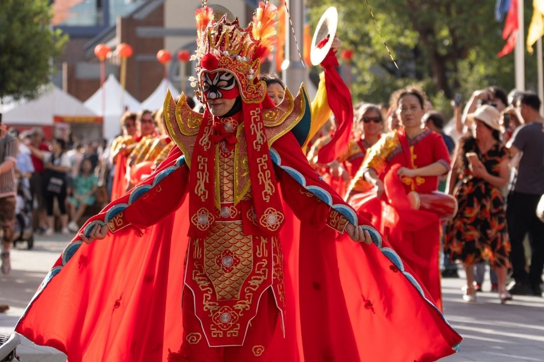 A vibrant parade scene with a person in an elaborate red and gold costume, wearing a feathered headdress, exuding joy and energy. Background shows more performers.