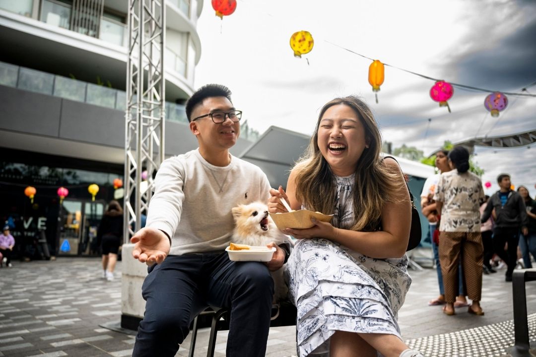 A joyful couple sits outdoors at a bustling street event, laughing. Colourful balloons float above, and people in the background enjoy the lively atmosphere.