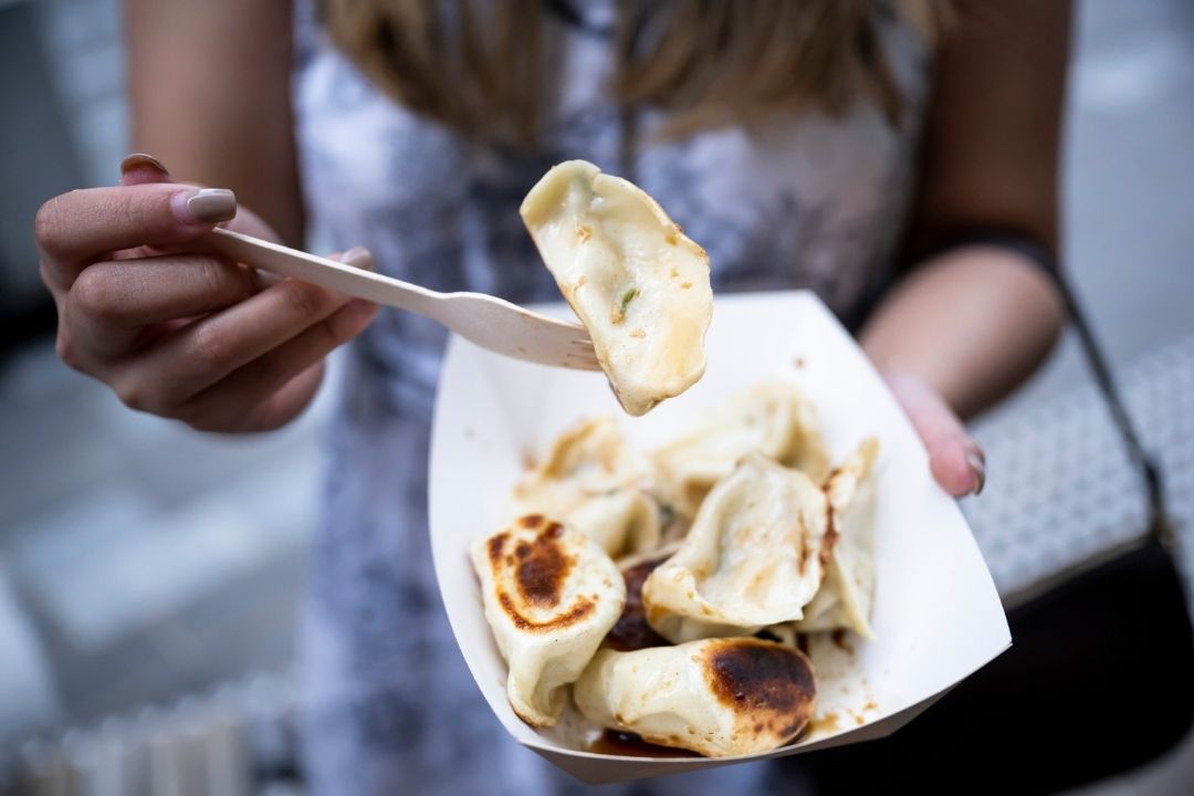 A person holds a tray of rolled crepes filled with berries and cream, using a wooden fork to pick one up. The scene conveys a sense of enjoyment and indulgence.