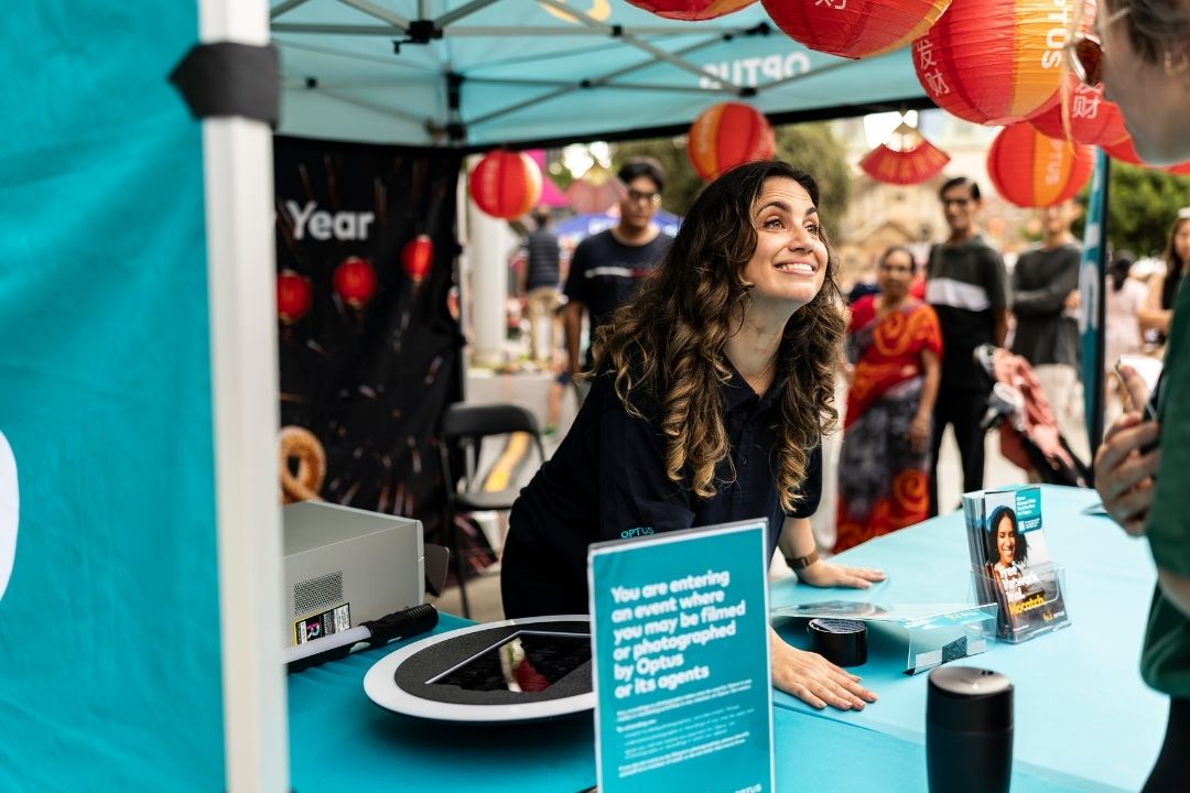 A woman smiles enthusiastically at an outdoor booth under a teal canopy. The table has a spinning wheel, promotional materials, and party balloons.