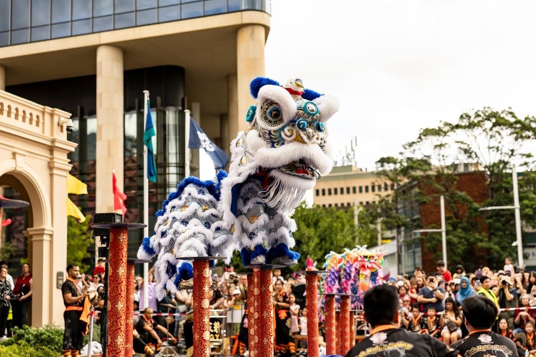 Lion dance performer on stilts in a vibrant white and blue costume captivates a large crowd in an outdoor urban setting, exuding excitement and festivity.