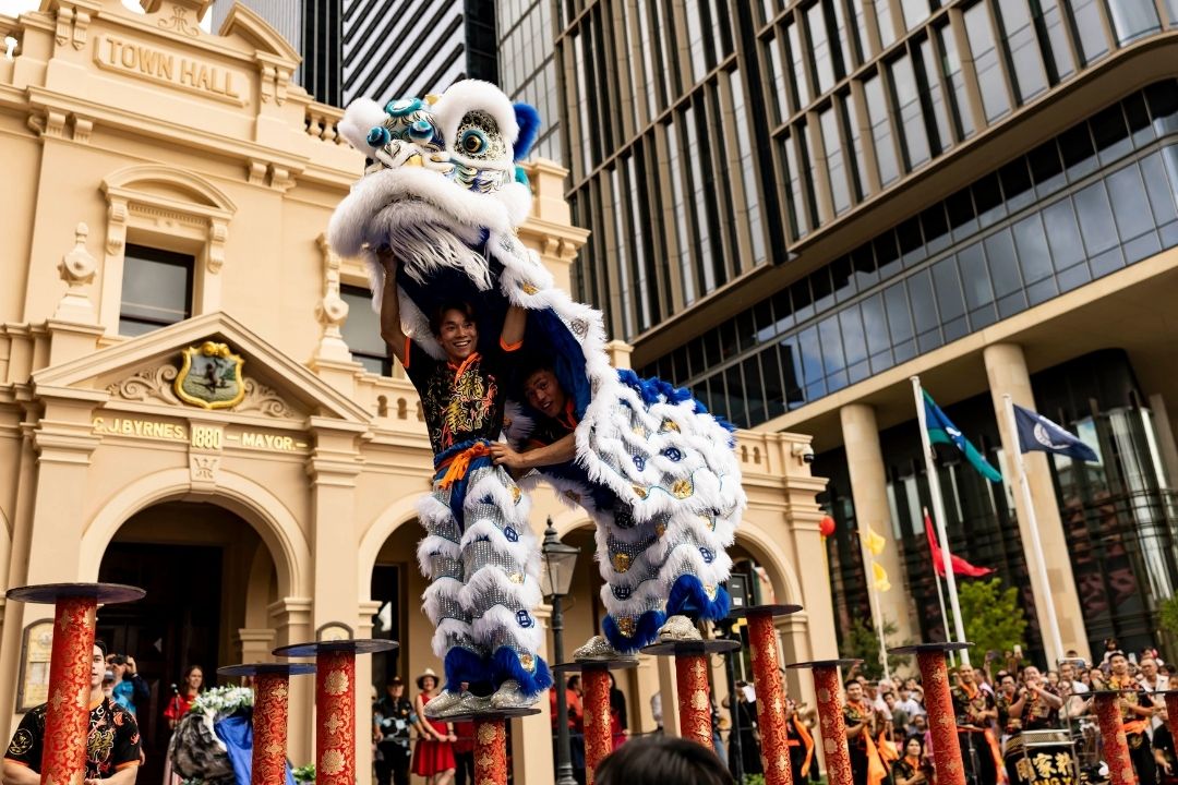 Lion dance performers in vibrant costumes balance on poles during a cultural festival. Historic and modern architecture form the backdrop, adding contrast.