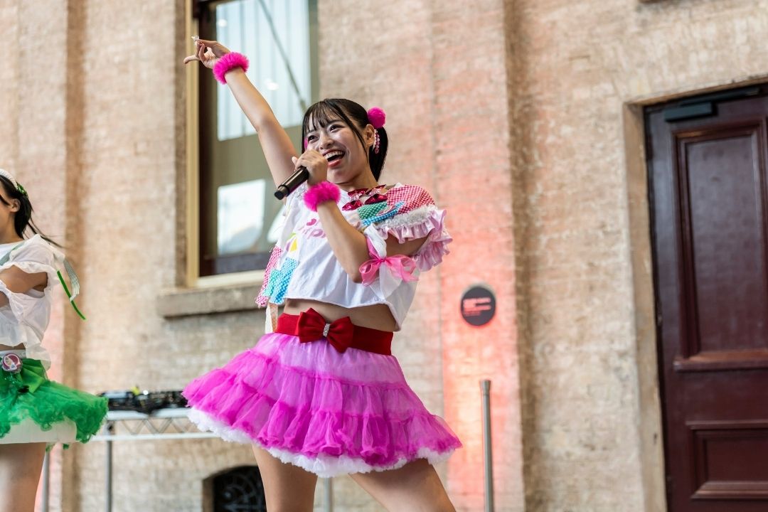 A performer in a vibrant pink and white outfit energetically sings on an outdoor stage. The scene conveys a lively and joyful atmosphere.