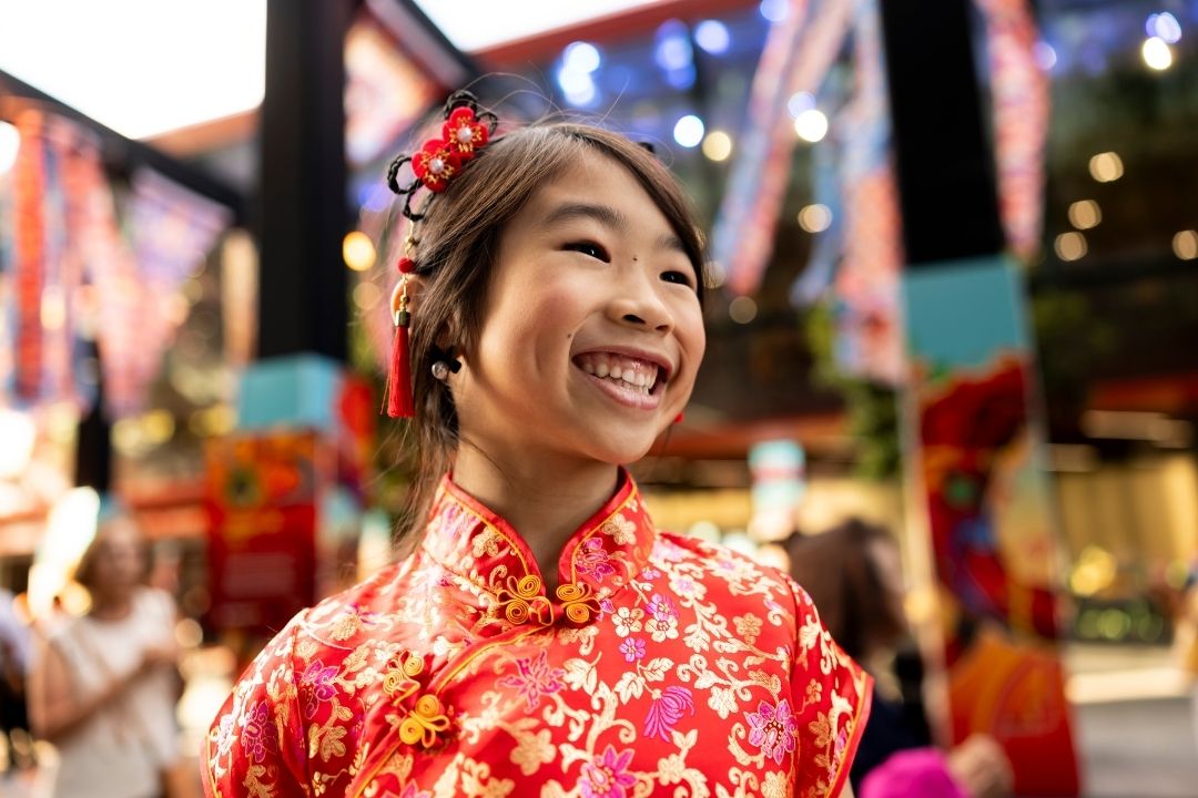 A woman in a vibrant floral dress with a flower in her hair smiles joyfully in a festive market setting, decorated with colourful lights and banners.