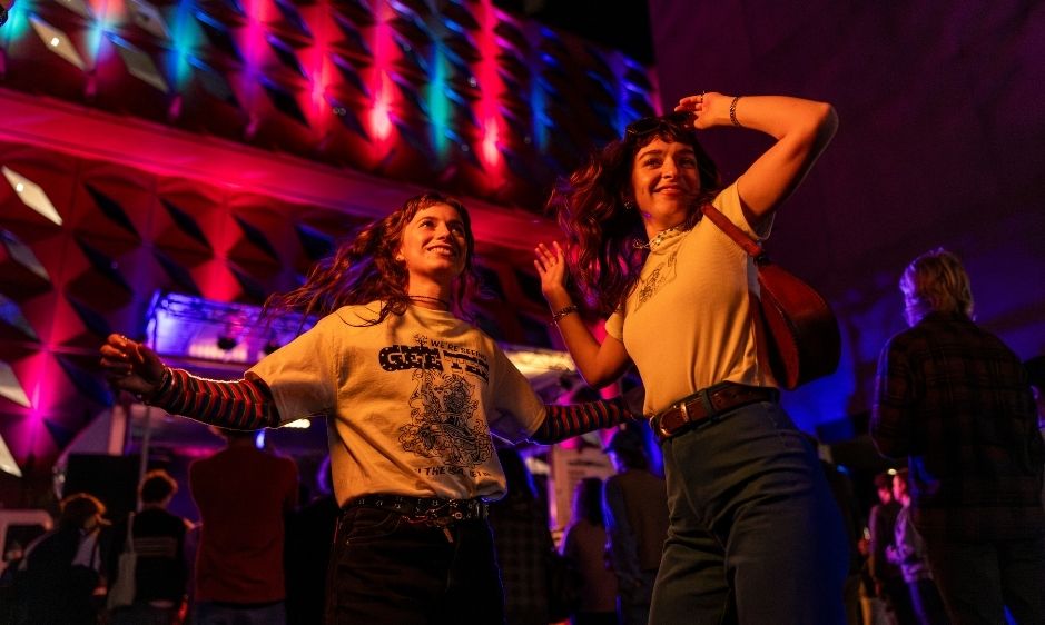Two people dance joyfully in a club with colourful lights illuminating the background. They wear casual clothes, expressing excitement and energy.