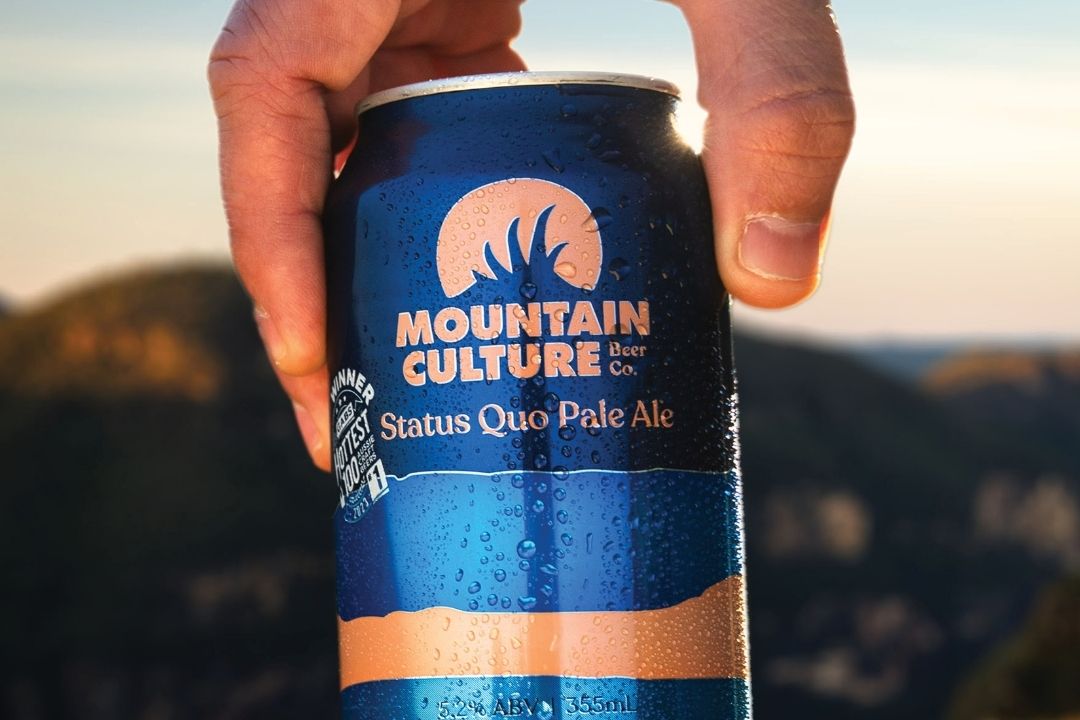 A hand holds a frosty can of Mountain Culture Status Quo Pale Ale against a blurred mountain backdrop, evoking refreshment and adventure.
