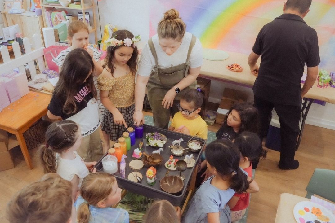 A diverse group of children and adults joyfully decorates cupcakes in a colorful room with a rainbow mural. The atmosphere is lively and creative.