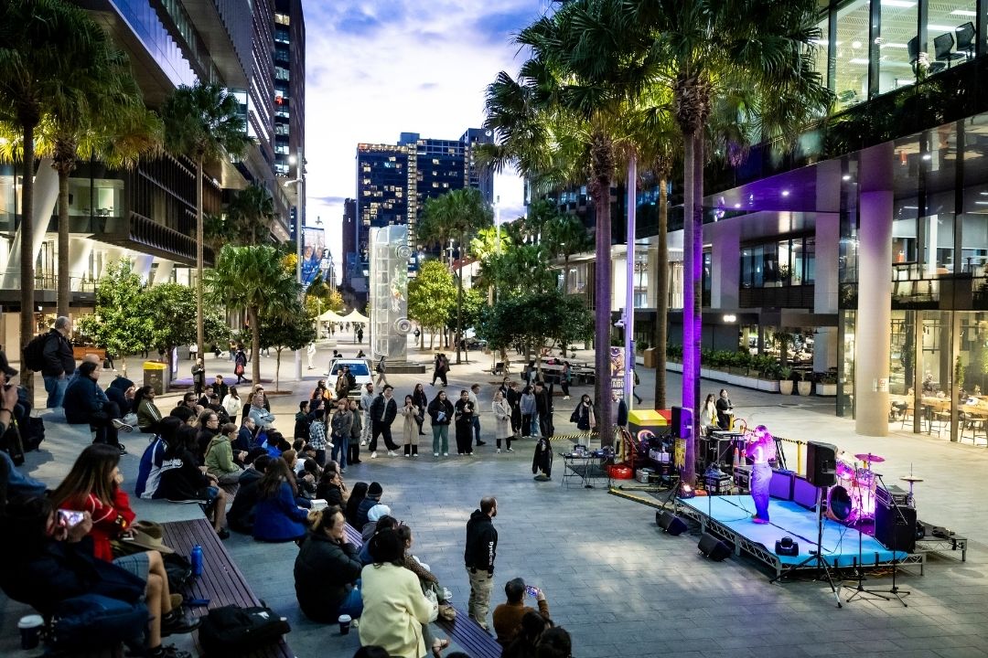 A lively street scene at dusk shows a crowd watching a live outdoor band performance. Tall buildings and palm trees frame the vibrant urban setting.