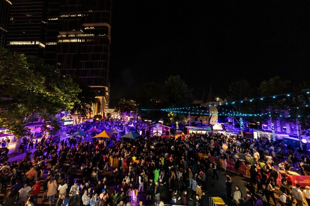 A vibrant street crowd gathered at night for a festival or celebration. The scene is illuminated by colourful lights, creating an energetic and festive atmosphere.