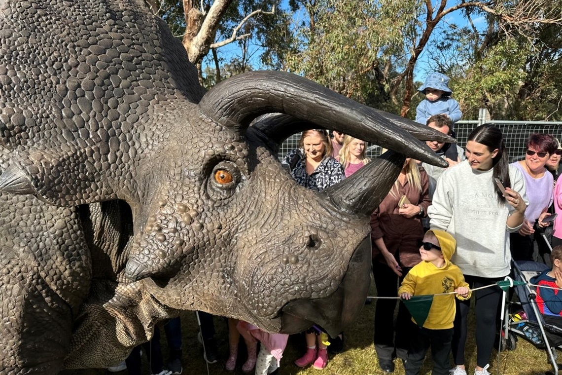Lifelike triceratops model in a crowded park setting, surrounded by curious children and adults. The scene is lively and filled with excitement.