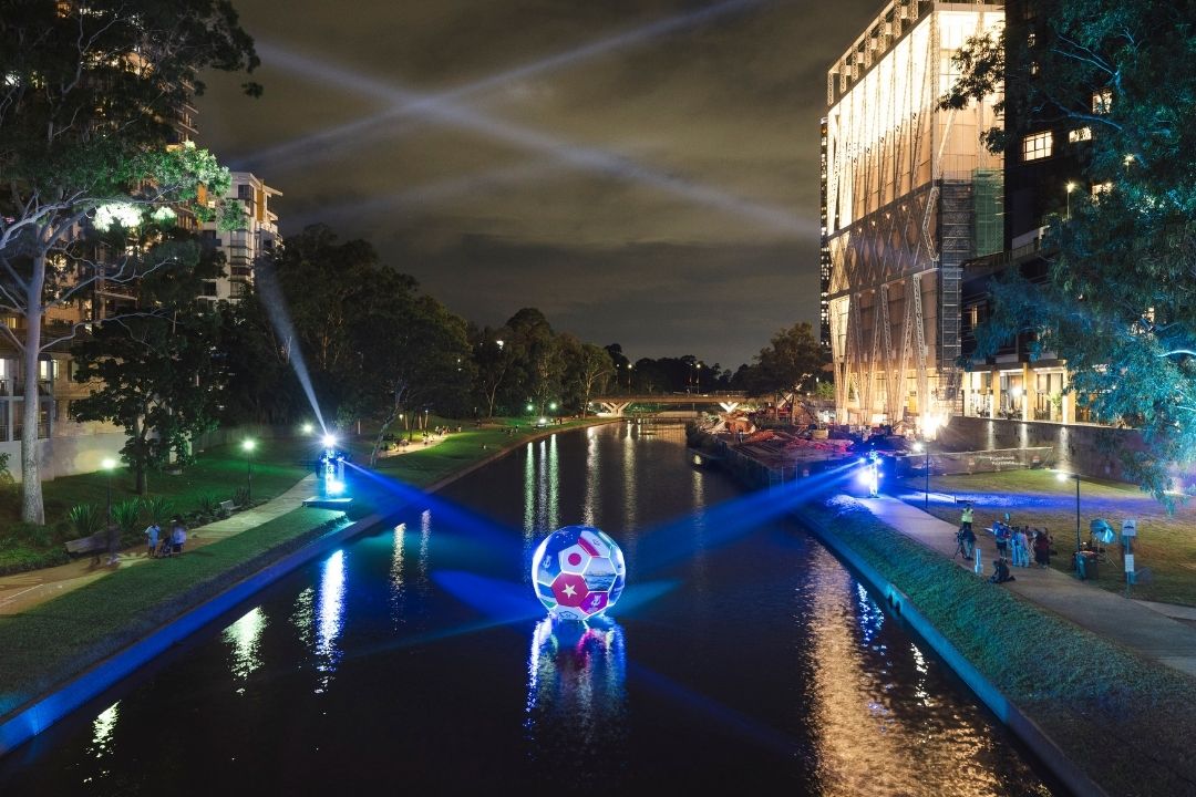 Night scene of an urban canal with colorful blue and white lights illuminating the water and surroundings, creating a serene, atmospheric ambiance.