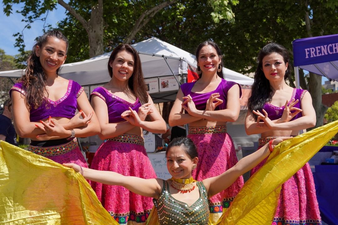 Five women in bright costumes, four in pink dresses and one in green, smile while posing outdoors at a festival, conveying joy and celebration.