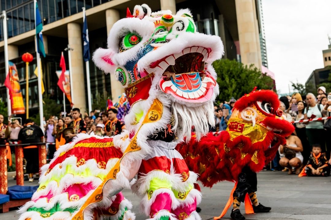 Three performers in colourful lion costumes are elevated on poles, with team members supporting them below. The scene is festive, set against a clear blue sky.