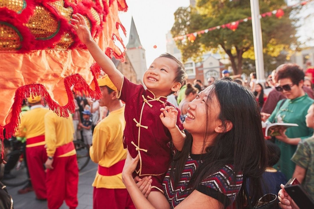 A child joyfully reaches toward a vibrant dragon costume during a cultural celebration, held by a smiling woman. Onlookers surround them, enjoying the festive parade.