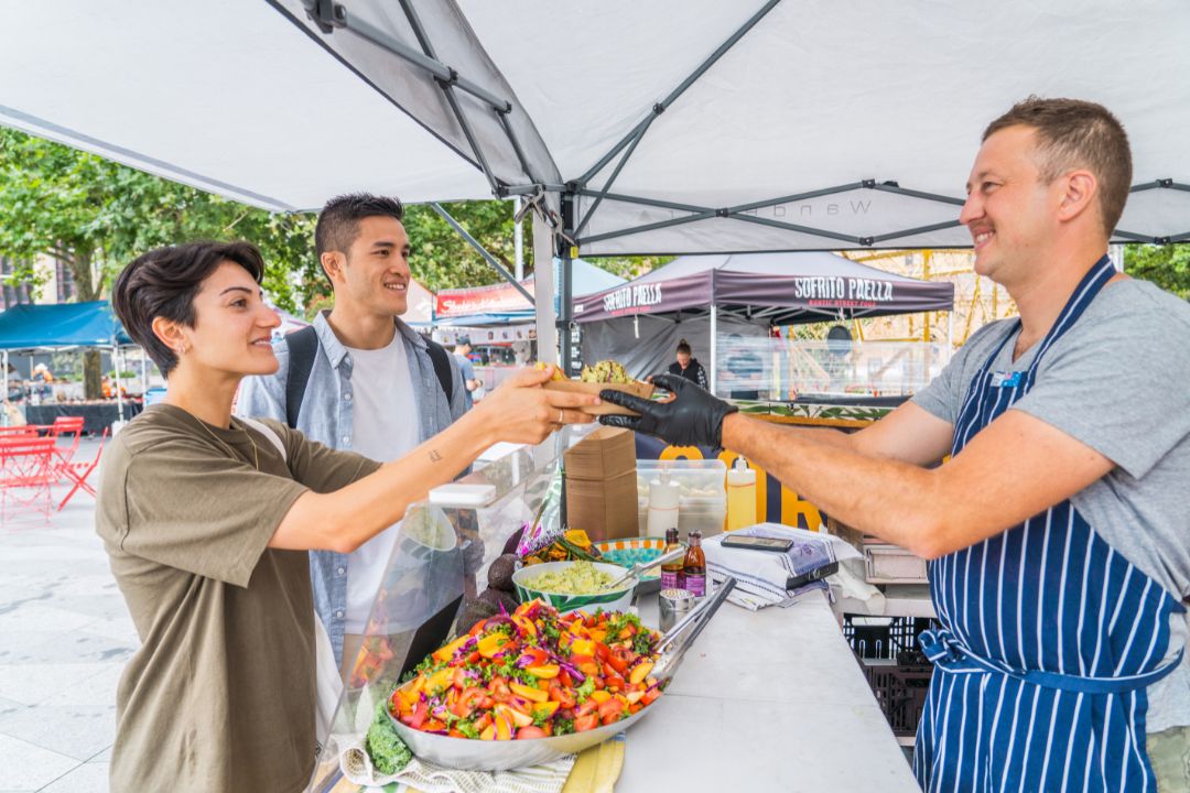 A smiling vendor in a striped apron serves food to two customers under a white tent at an outdoor market. The atmosphere is cheerful and lively.