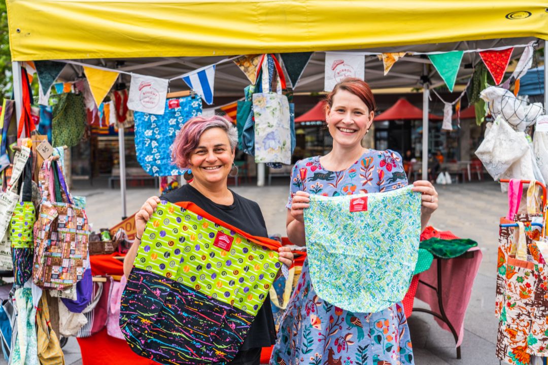 Two smiling women hold colorful garments under a yellow canopy at an outdoor market. Brightly patterned fabrics hang around them, creating a cheerful, lively scene.
