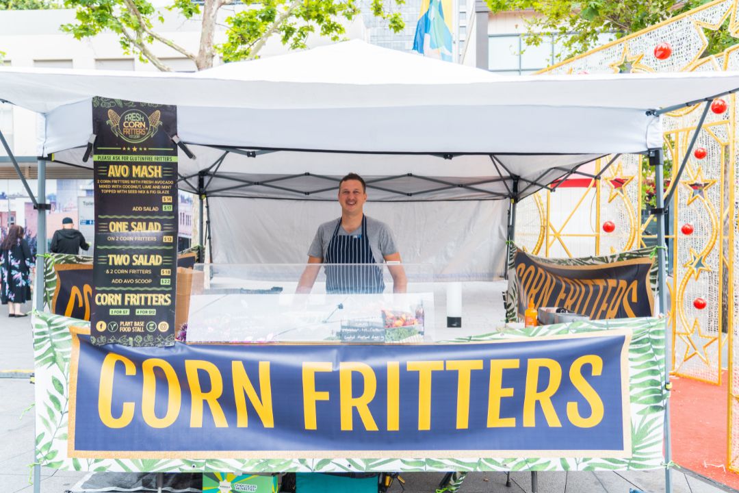 A vendor stands smiling at a "Corn Fritters" stall under a white canopy at an outdoor market, with vibrant banners and a menu displayed.