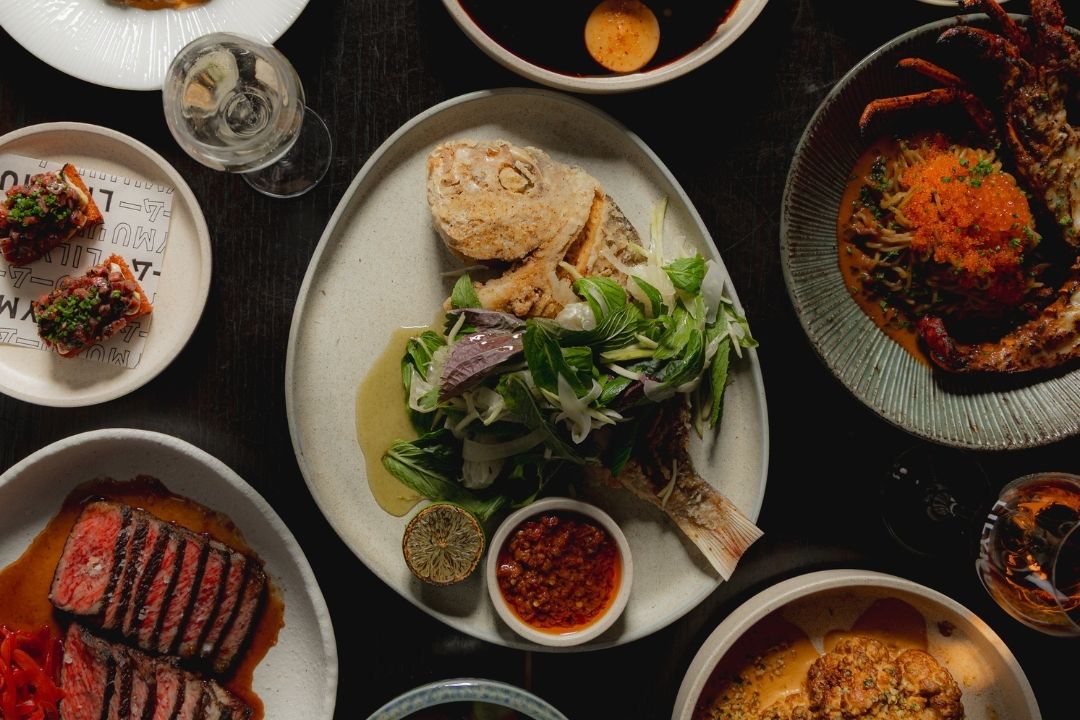 A variety of elegantly plated dishes on a dark table: grilled steak, roast chicken with greens, pasta, and desserts, creating a warm, inviting dining experience.