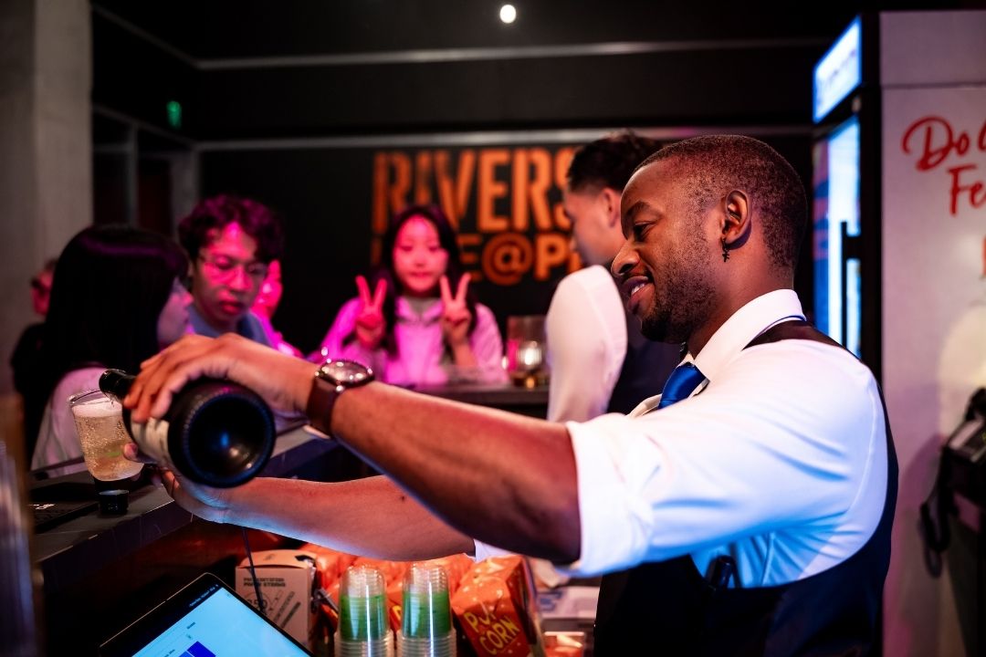 A bartender in a white shirt pours a drink at a lively bar. People in the background chat and smile, creating a festive and vibrant atmosphere.