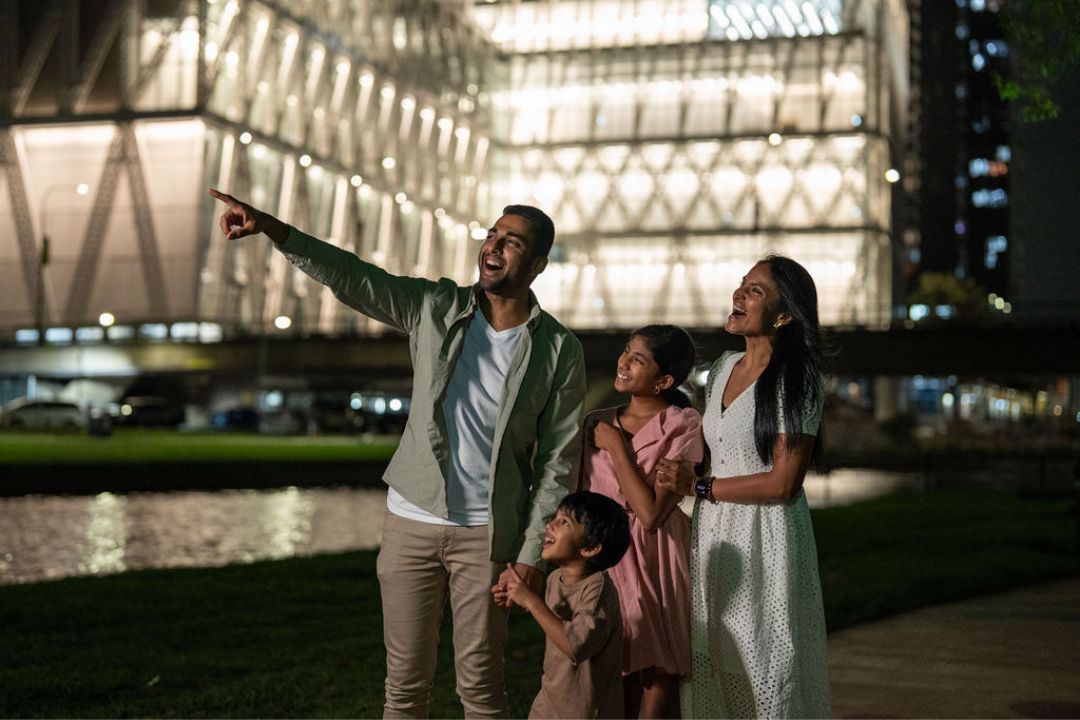 A family of four stands together, smiling and pointing at a brightly lit modern building at night. They seem joyful, enjoying a night out by a waterway.