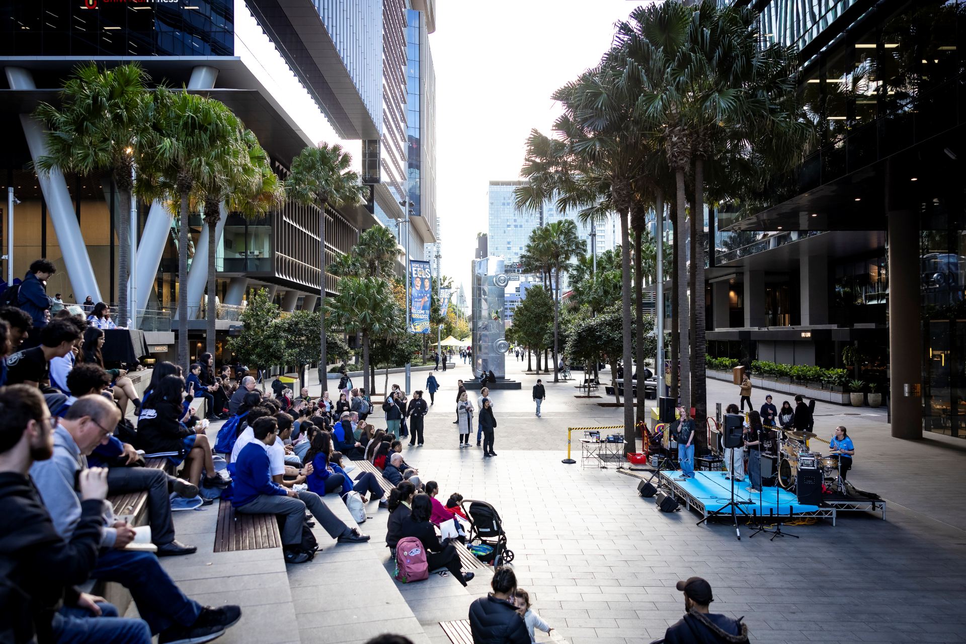 A bustling urban plaza with many people seated and walking, surrounded by tall buildings and palm trees, creating a vibrant, lively atmosphere.