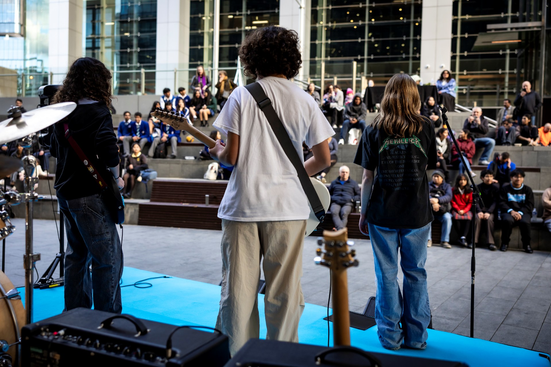 Three musicians perform on an outdoor stage, facing a large, seated audience. The lively atmosphere suggests enjoyment and engagement.