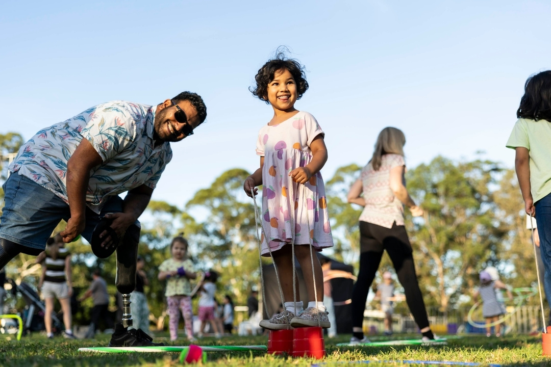 A joyful child in a pink dress stands on grass, holding a red bucket, flanked by smiling adults on a sunny day in the park. Trees and clear sky in background.